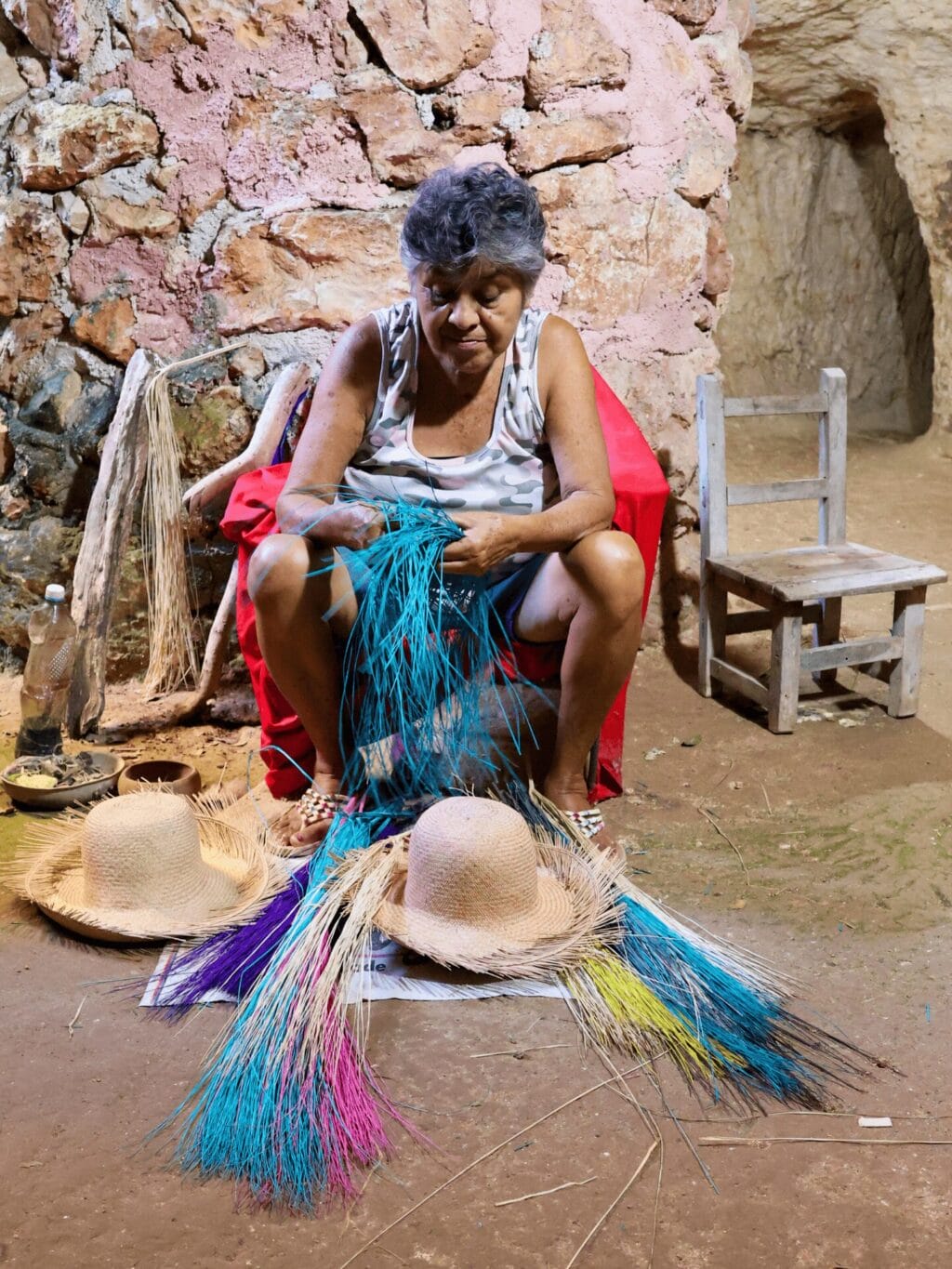 Eine mexikanische Frau mit schwarzen, graumelierten Haaren, sitzt in einer Kellerhöhle auf einem Stuhl beim Flechten eines türkisfarbenen Huts. Am Boden vor ihren Füssen liegen zwei fast fertig geflochtene, naturfarbene Strohhüte sowie farbige Strohhalme.